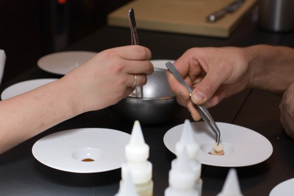 Two chefs lean over ingredients, plating with tweezers.