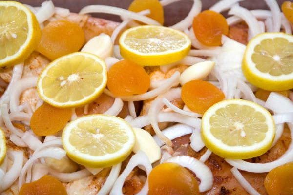 Chicken in roasting pan, skin side up, topped with onions, garlic cloves, apricots and lemon slices.